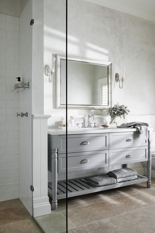 Bright, refined traditional bathroom featuring a glass-enclosed tiled shower and a freestanding grey vanity with four drawers, open slatted lower shelf, white countertop, and chrome fixtures. A large framed mirror, paired wall sconces, white wall panelling, and neutral stone-look floor tiles create a classic, softly layered interior.