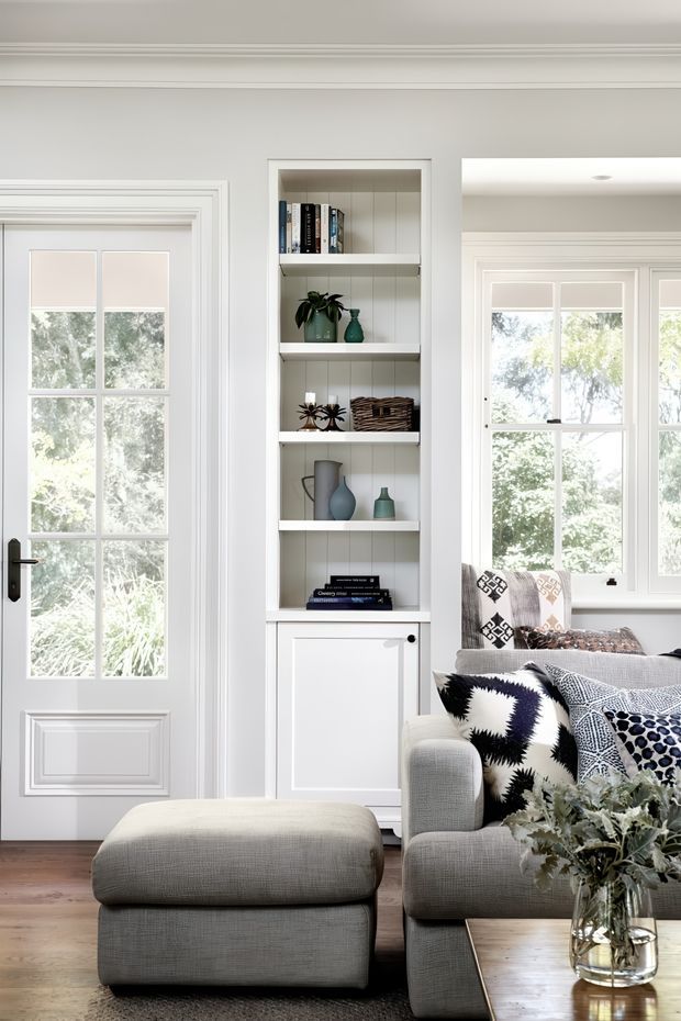 Bright, refined living room with white paneled walls, detailed mouldings, a glazed French-style door, built-in open shelving, and large divided-light windows. A grey upholstered sofa and ottoman sit around a timber coffee table, accented by monochrome patterned cushions and neutral decorative objects. The room has a calm, polished, traditional-meets-contemporary appearance.