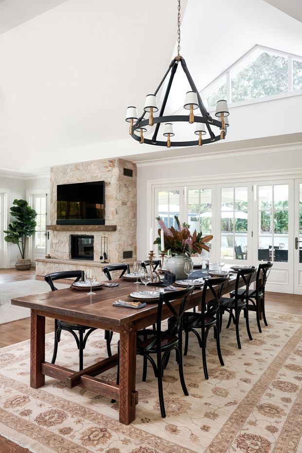 A bright, refined dining room with a large rustic dark-wood dining table set for eight, black cross-back chairs, and a patterned area rug. A substantial black-and-brass ring chandelier hangs from the high sloped ceiling. The room includes a stone-clad fireplace wall with a mounted television, white built-in trim and French doors opening toward a light-filled outdoor area. Large clerestory glazing and pale walls create an airy, elegant atmosphere.