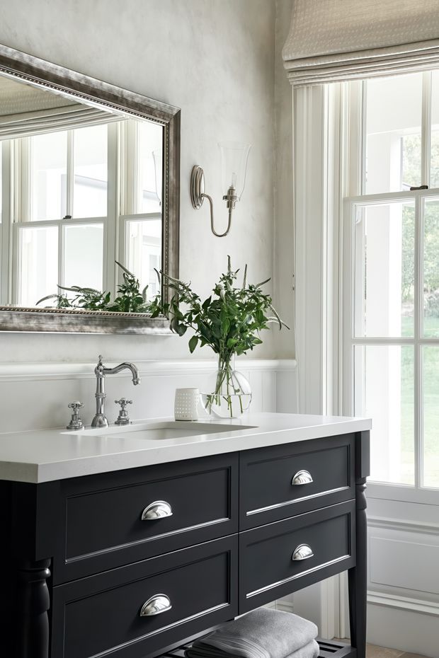 Elegant traditional bathroom vanity area with a dark painted double-drawer vanity, white countertop and integrated basin, polished chrome plumbing fixtures, wall panelling, a large framed mirror, and tall white-framed windows dressed with Roman blinds. A leafy floral arrangement adds natural softness.
