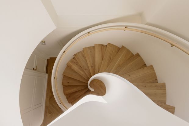 Top-down view of an elegant curved spiral staircase with warm timber treads, white sculptural balustrades and curved handrails. The staircase is set within a bright, refined interior with white paneled walls and ceiling details.
