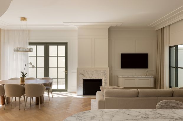 Elegant open-plan living and dining room with a warm neutral palette, herringbone timber flooring, extensive white wall and ceiling mouldings, a marble fireplace, built-in TV cabinetry, tall French doors, and full-height curtains. A dining table occupies the left side while a large upholstered sofa and marble-topped coffee table define the living area.