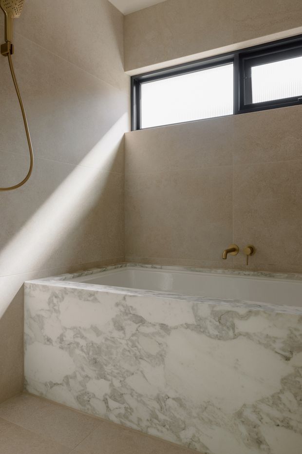 A contemporary bathroom featuring a built-in white bathtub clad in bookmatched-look veined marble or marble-effect stone, beige wall and floor tiles, and brushed brass/gold tapware. A high horizontal black-framed window provides natural light, creating a warm, refined atmosphere.