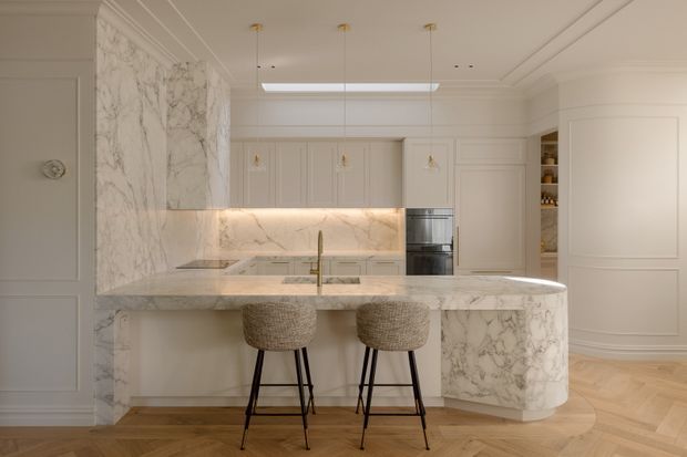 Elegant, warm-toned kitchen featuring extensive white veined marble on the island and backsplash, pale cabinetry with decorative panel detailing, integrated appliances, brass tapware, pendant lighting, and two woven bar stools. The curved island edge and herringbone timber flooring add softness and visual interest, while the coffered and moulded ceiling contributes a refined classic character.