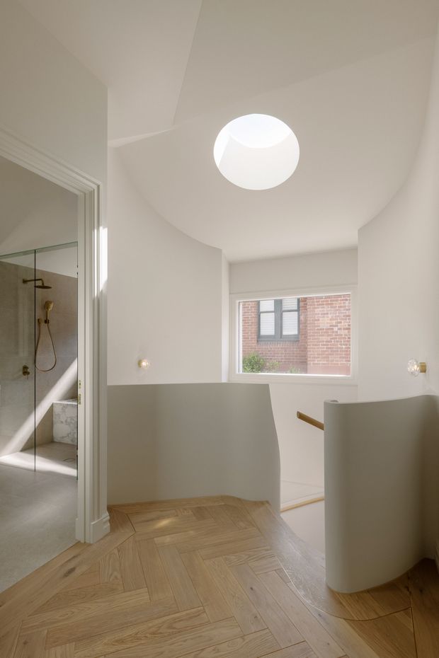 A bright, sculptural bathroom interior with curved white walls and half-height partitions, a circular skylight, and a large window overlooking a brick building. The foreground features warm light timber herringbone flooring and a curved white freestanding or built-in vanity partition. A glass-enclosed shower with brass-toned fittings is visible to the left, while a stair or lowered bathroom zone appears beyond the curved wall.