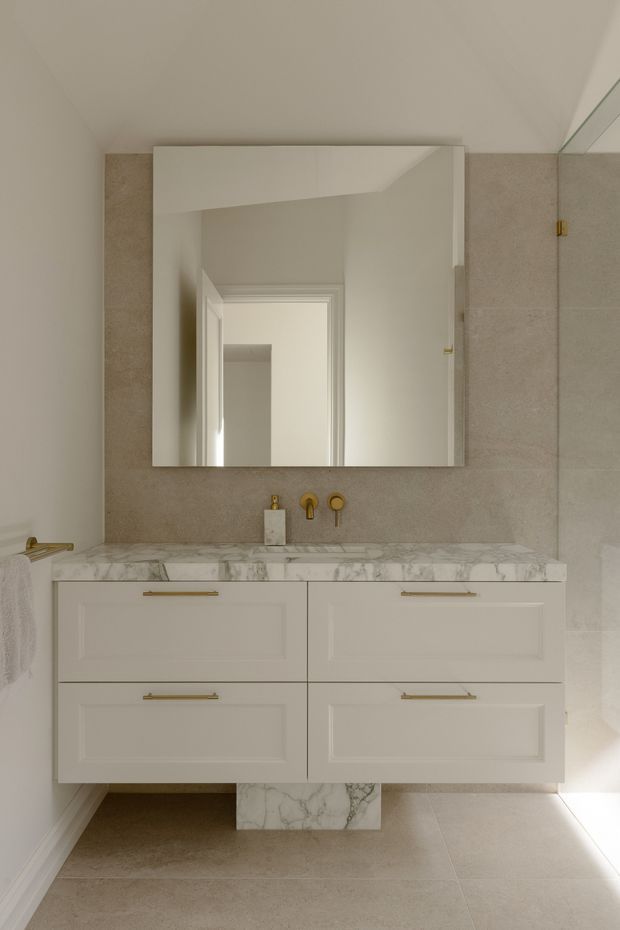 A refined bathroom vanity area with a wall-mounted white shaker-style double vanity, veined white marble benchtop, brass tapware and handles, a large frameless mirror, and large warm beige stone-look wall and floor tiles. The space is softly lit and understated, with a small towel rail visible at the side.
