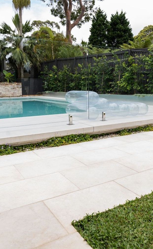 A landscaped outdoor pool area with a turquoise swimming pool, pale stone or concrete paving, a transparent glass safety barrier, low planting, and a dark timber privacy fence backed by tropical and mature greenery. The clean-lined pool edge and expansive light paving create a contemporary residential setting.