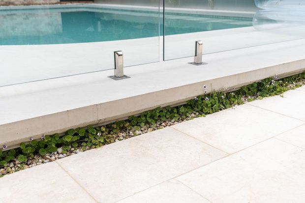 Close-up exterior view of a modern swimming pool edge with a clear glass safety balustrade supported by stainless-steel posts. A pale concrete coping and large light paving slabs border the pool, with a narrow planted drainage strip of low greenery and pebbles beneath the raised edge.