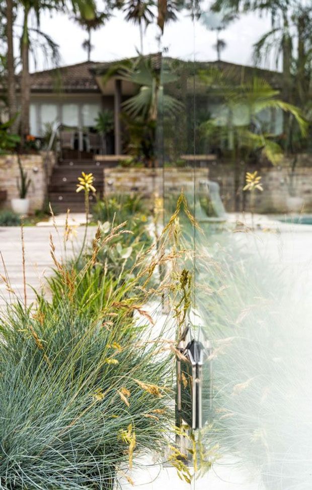 Blurred exterior view of a landscaped residential pool area, with ornamental grasses and planting in the foreground, a pale pool surface at right, and a low stone-clad house with broad glazing and a tiled roof in the background. A dark-framed glass pool fence and small garden light are visible.