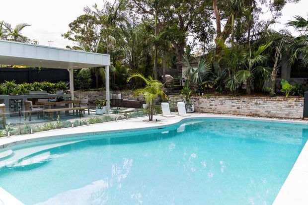 A bright outdoor pool area with a large turquoise swimming pool in the foreground, surrounded by white pool paving, a raised pale stone retaining wall, lush tropical planting, and a covered outdoor dining and entertaining pavilion with glass screening and timber furniture.