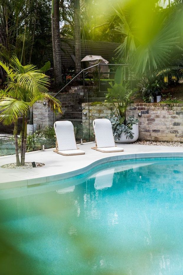 A tropical landscaped pool area with a curved turquoise swimming pool, pale concrete surrounds, two white loungers, lush palms and planting, raised weathered brick retaining walls, and a stairway leading into dense greenery.
