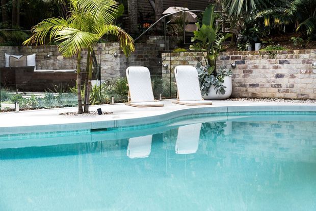 A landscaped outdoor swimming pool with clear turquoise water, a pale paved pool surround, two white woven loungers, tropical palms and planting, and a weathered brick retaining wall in the background. The scene has a relaxed resort-like atmosphere.