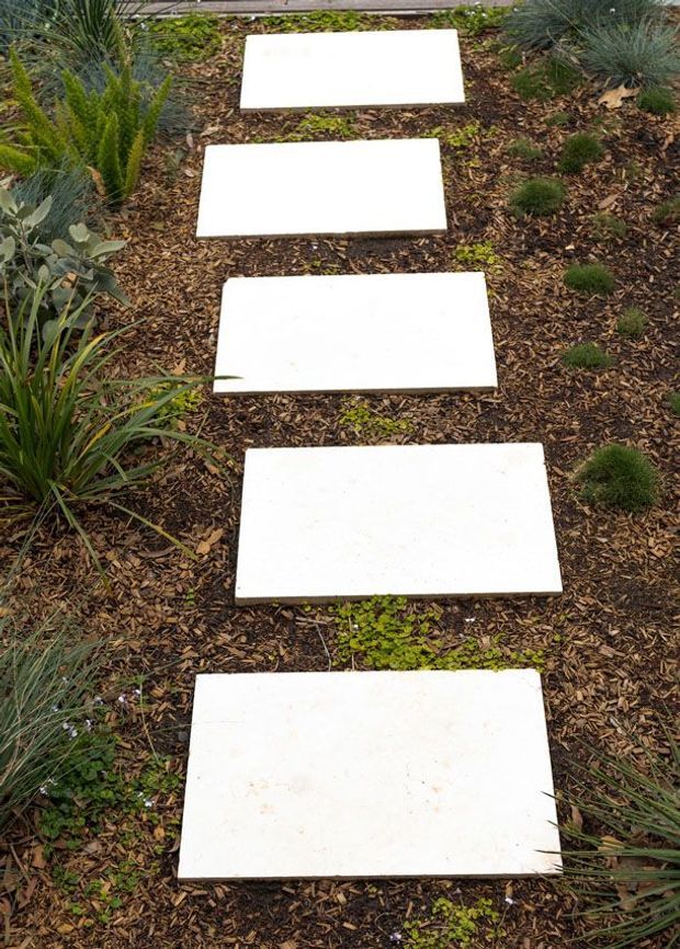 A landscaped garden path made from a series of large rectangular white stepping stones set into dark brown mulch. Low ornamental grasses, succulents, and other drought-tolerant planting border the path, creating a naturalistic outdoor walkway.