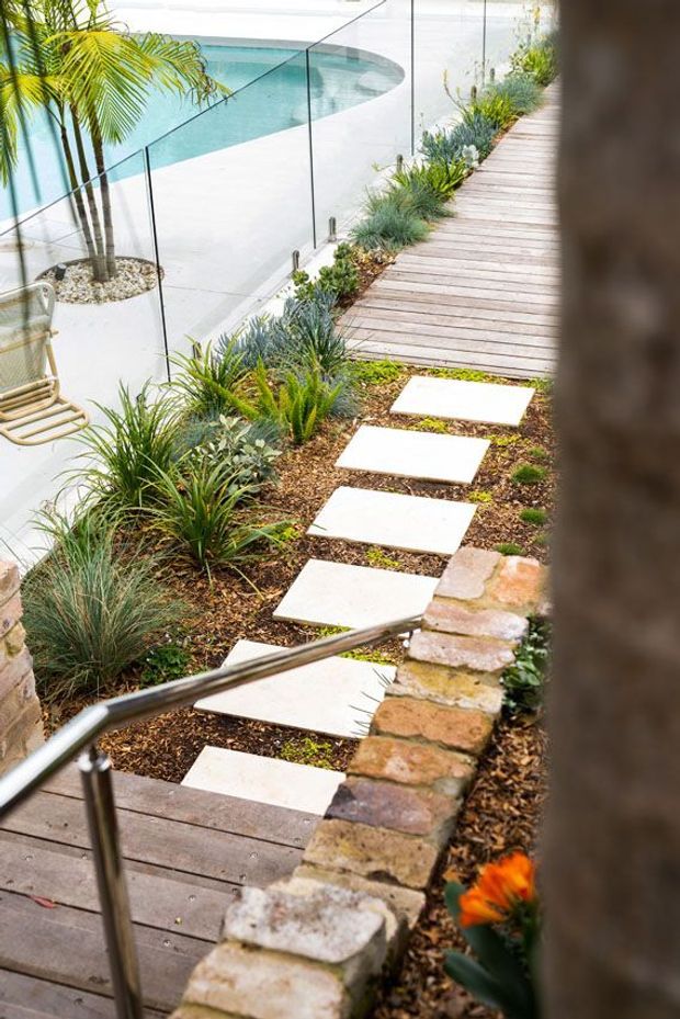 A landscaped outdoor poolside area viewed from above, with a timber boardwalk and steps, pale rectangular stepping stones through a planted garden bed, low brick retaining walls, stainless steel handrails, and frameless glass pool fencing. A curved turquoise swimming pool and palms are visible beyond.