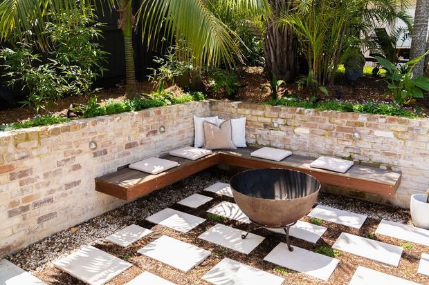 A lush tropical courtyard lounge with a curved exposed-brick retaining wall, built-in timber bench seating, scattered light stepping pavers over gravel, and a central round metal fire bowl. Dense palms and tropical planting create a private outdoor setting.
