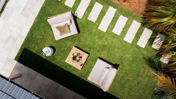 Aerial view of a landscaped outdoor courtyard or garden area with a rectangular lawn, pale stepping-stone path, paved perimeter, two light upholstered outdoor lounge chairs, and a central wooden fire pit or low table. Palm plants border the lawn, creating a resort-like outdoor setting.