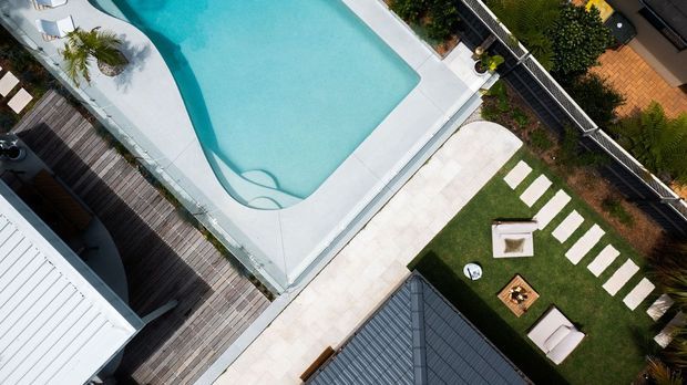 Aerial view of a contemporary residential outdoor area featuring a large turquoise swimming pool with curved edges, pale paving, timber decking, and a landscaped lawn with stepping stones and outdoor seating.