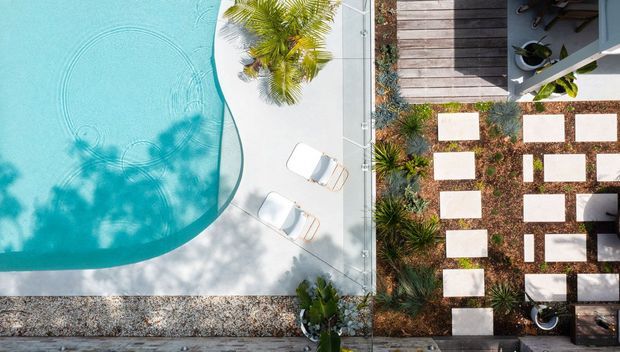 Aerial view of a contemporary outdoor property showing a curving turquoise swimming pool beside a pale paved pool terrace, white sun loungers, tropical planting, and a structured garden path of rectangular stepping stones through dark planting beds. A timber deck with outdoor furniture occupies the upper-right area.