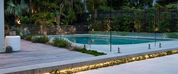 A landscaped outdoor pool area viewed from a paved terrace, featuring a turquoise swimming pool enclosed by frameless glass balustrades, timber decking, integrated linear lighting, tropical planting, and a dark privacy fence with a low masonry retaining wall.