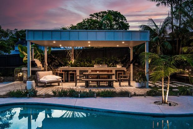 A tropical outdoor entertaining area beside a swimming pool, featuring a covered pavilion with a blue metal fascia, timber posts, built-in outdoor kitchen cabinetry and benchtop, a long dining table with benches, lounge seating, and lush palm landscaping at dusk.
