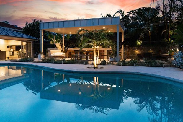 Twilight view of a landscaped residential pool area with a large blue swimming pool in the foreground, a covered outdoor entertaining pavilion with a flat roof and illuminated ceiling, timber seating and furnishings, tropical palms, and a partially visible house with a tiled roof at left. The pool reflects the pavilion and evening sky.