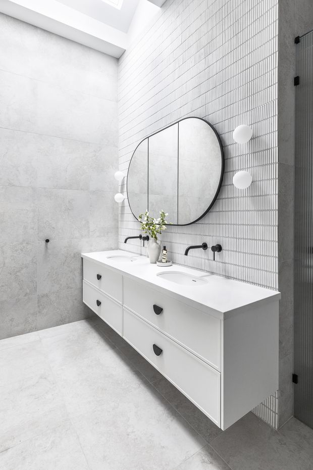 Bright contemporary bathroom featuring a floating double vanity with white drawer cabinetry and integrated white basins, black wall-mounted taps, a large rounded black-framed mirror, globe wall lights, pale grey stone-look wall and floor tiles, and a vertically ribbed feature wall. A skylight adds abundant natural light.