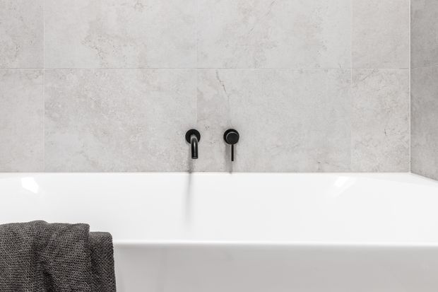 Close-up view of a minimalist bathroom bathtub with a white rectangular bath, wall-mounted matte black tapware, and large-format light grey stone-look wall tiles. A dark grey towel is draped over the bath edge.