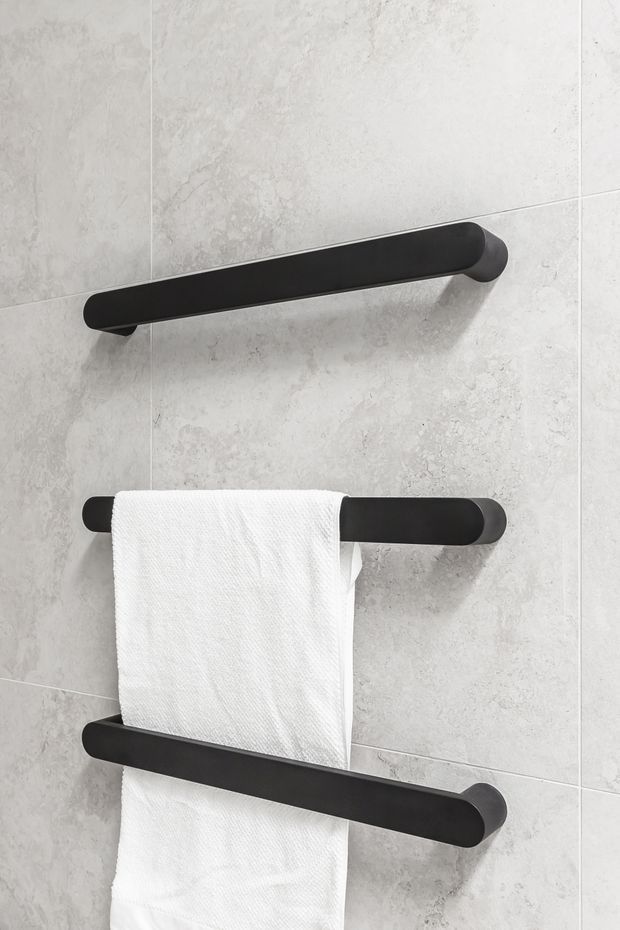 Close-up of three matte black horizontal towel rails mounted on a light grey textured tiled wall, with a white towel hanging from the middle rail. The image focuses on contemporary bathroom hardware and finishes.