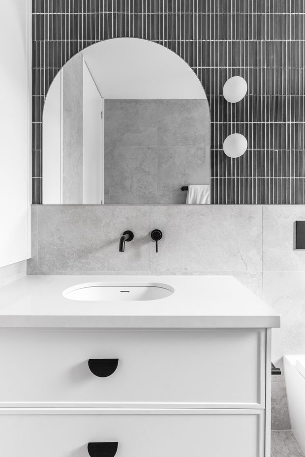 A monochrome contemporary bathroom vanity with a white integrated basin, pale grey tiled wall and splashback, matte black wall-mounted tapware, and a large arched mirror. The mirror reflects the tiled shower area, while a black linear tile feature wall and two round wall lights add graphic contrast. White built-in vanity drawers occupy the foreground.