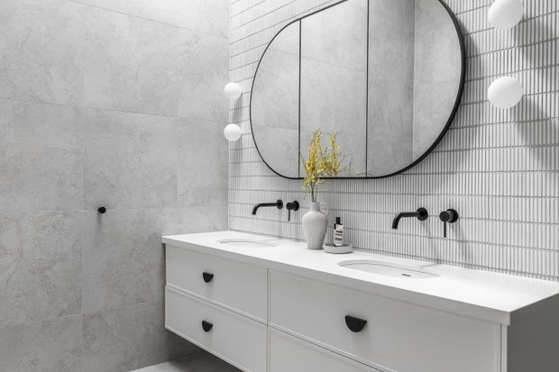 Contemporary double-vanity bathroom with a long white floating cabinet, integrated basins, black wall-mounted tapware, pale grey large-format wall tiles, and a vertically ribbed white tiled feature wall. A large rounded rectangular black-framed mirror and spherical wall lights create a refined monochrome look, with a small vase providing a yellow accent.