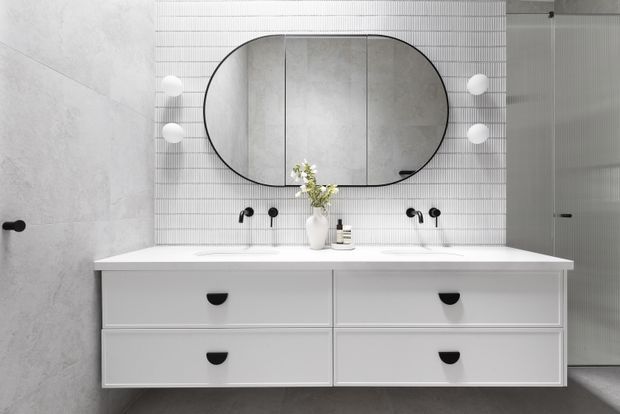 Contemporary minimalist bathroom featuring a wide double vanity with white drawers, a white benchtop, twin black wall-mounted tap and basin fixtures, and a large rounded-rectangle mirror. The vanity wall is finished in white narrow vertical tiles, contrasted by pale grey stone-look wall and floor finishes. Black hardware and fittings provide restrained contrast, with a glass shower enclosure visible at right.