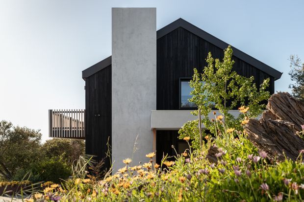 Exterior view of a contemporary dark-clad gable-roof house, featuring vertical black timber-like cladding, a tall smooth concrete chimney or sculptural wall, a projecting balcony, and simple dark-framed windows. The building is partially screened by a naturalistic flowering garden and rocky landscaping.
