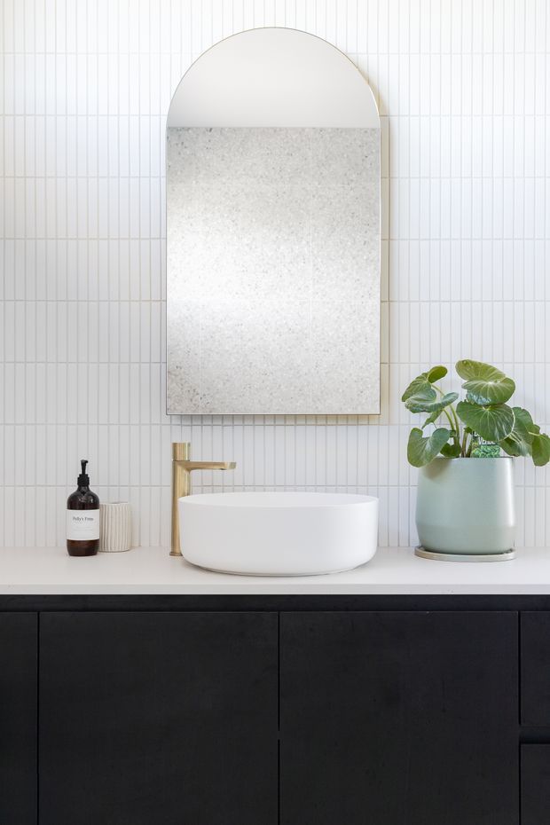 A bright, refined bathroom vanity featuring a white vessel basin on a pale benchtop, a brushed brass wall-mounted tap, dark charcoal cabinetry, and a large arched mirror. The wall is finished with narrow vertical white tiles, complemented by a soft green potted plant and minimal toiletries.