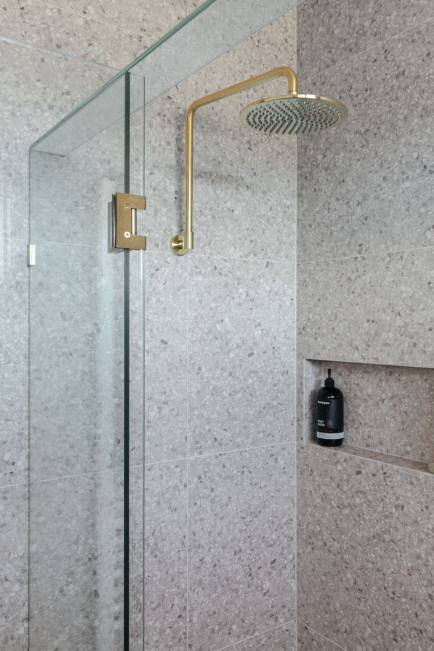 Close-up view of a contemporary walk-in shower enclosed by clear frameless glass, with large-format grey terrazzo-look wall tiles, a recessed shower niche, and a brushed brass rainfall showerhead and controls. A black bottle sits in the niche.