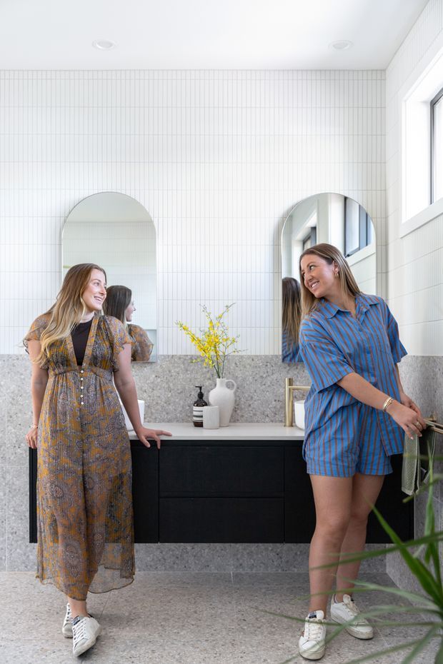 Bright contemporary bathroom featuring a full-width grey terrazzo vanity backsplash and floor, matte black floating cabinetry, white vertically lined wall tiles, arched mirrors, and a light stone benchtop. Two people are present, with natural light entering through a side window and a small plant in the foreground.