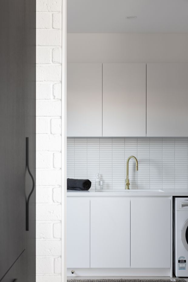 A compact, minimalist laundry room viewed through a doorway. The space features white flat-panel cabinetry, a white tiled splashback with narrow horizontal tiles, a white benchtop and integrated sink, a brass gooseneck tap, and a front-loading washing machine. A white-painted brick wall and dark timber door frame the room.
