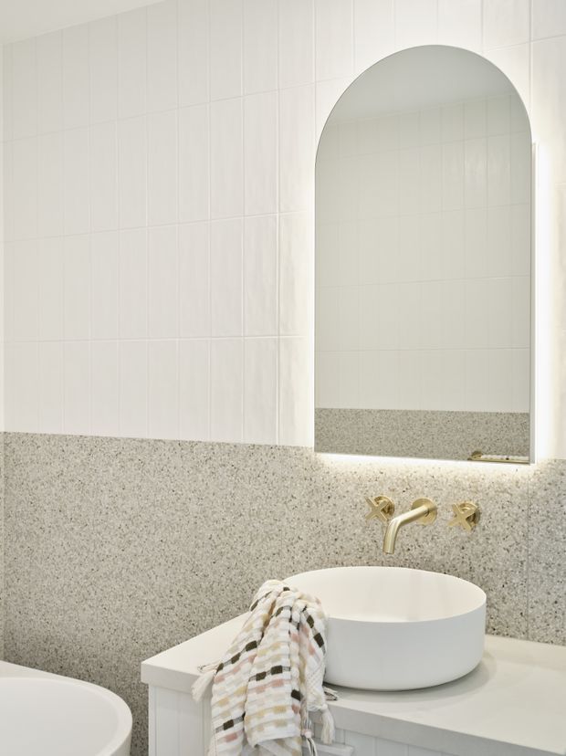 Bright contemporary bathroom vanity area featuring white rectangular wall tiles, a speckled terrazzo-like wall and benchtop, a white round vessel basin, brushed gold wall-mounted tapware, and a tall arched mirror with concealed under-mirror lighting. Part of a freestanding bathtub is visible in the foreground.