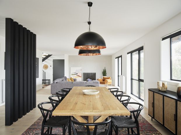 A bright, open-plan dining and living area featuring a large natural timber dining table surrounded by black dining chairs, a sculptural black pendant light with copper-toned interior, pale flooring and walls, and full-height black-framed glazing along one side. A black vertical-slat room divider separates the dining zone from the living area, which includes a grey sofa and warm-toned armchairs. A dark woven-front sideboard and patterned rug add contrast.