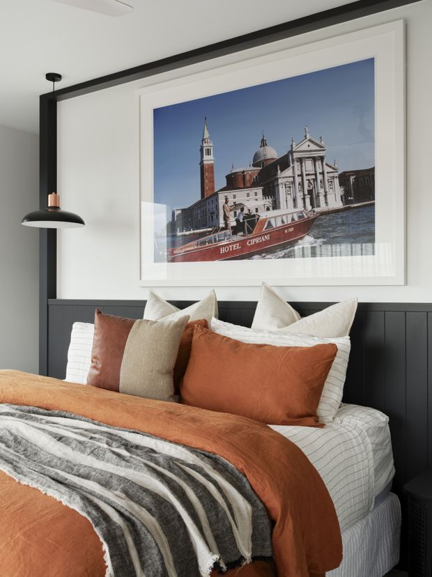 A styled bedroom with a layered bed dressed in rust-orange and neutral bedding, textured cushions, and a striped throw. The room features a dark vertical-panel headboard wall, white walls and trim, a black pendant light with a copper accent, and a large framed Venice architectural photograph above the bed.
