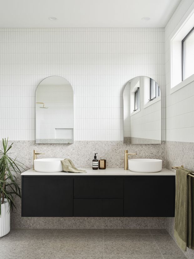 Bright contemporary bathroom featuring a floating matte-black double vanity with two white vessel basins, brass wall-mounted taps, arched mirrors, a terrazzo-look grey wall and floor finish, and white vertically lined wall tiles. A large window provides natural light, with a plant and folded towels adding softness.