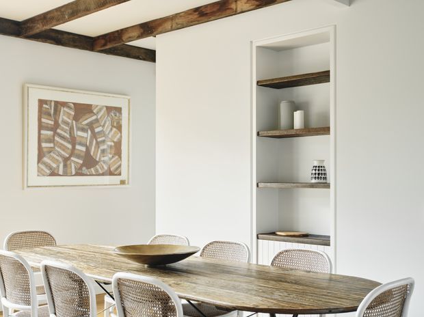 Bright dining room with a long rustic timber dining table surrounded by woven cane-back chairs. White walls and built-in open shelving display simple neutral ceramics, while exposed dark timber ceiling beams and framed abstract artwork add warmth and texture.