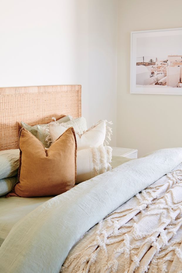 Bright, softly styled bedroom with a woven rattan headboard, layered neutral and tan pillows, pale bedding, and a textured cream throw. A framed coastal architectural photograph hangs on the light neutral wall, reinforcing a relaxed natural aesthetic.