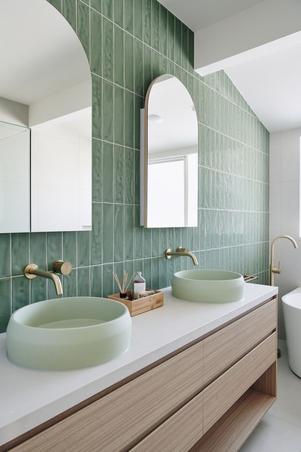 Bright contemporary bathroom featuring a long timber-look double vanity with two pale green vessel basins, brushed brass wall-mounted taps, and a large arched mirror. The walls are clad in glossy vertically oriented green tiles, with white tiled surfaces and a glass shower enclosure visible at the side.