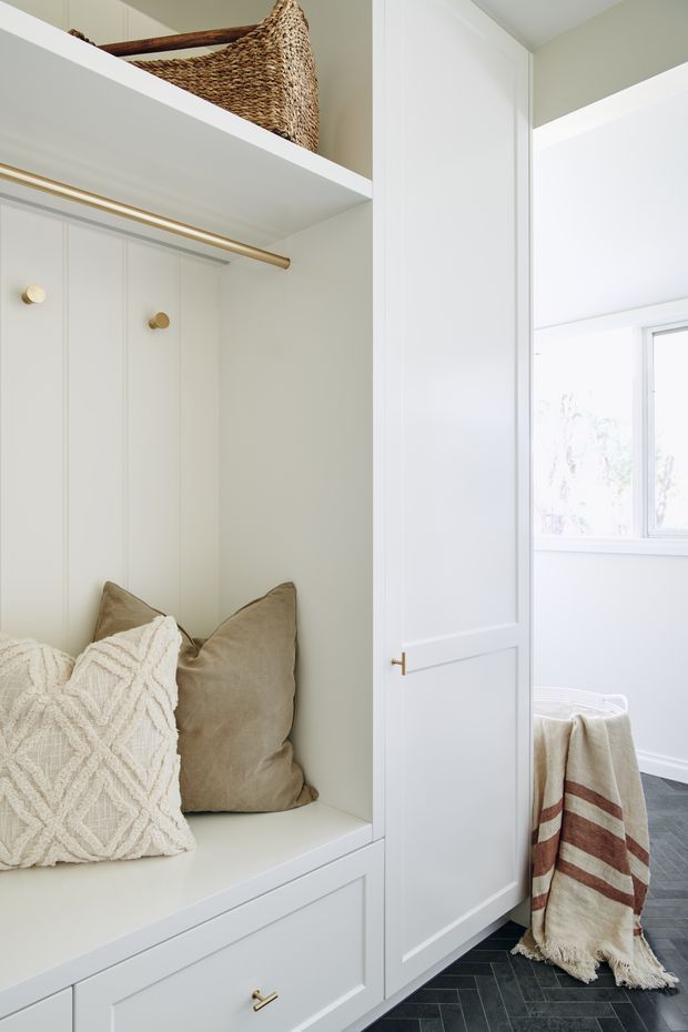 Bright, compact built-in dressing or entry storage nook with white paneled cabinetry, an integrated cushioned bench, open shelving, and a brass hanging rail. Woven baskets and neutral cushions add warmth. A partially visible adjacent room has a window, white wall moulding, and a dark herringbone floor, with a striped throw draped over a small piece of furniture.