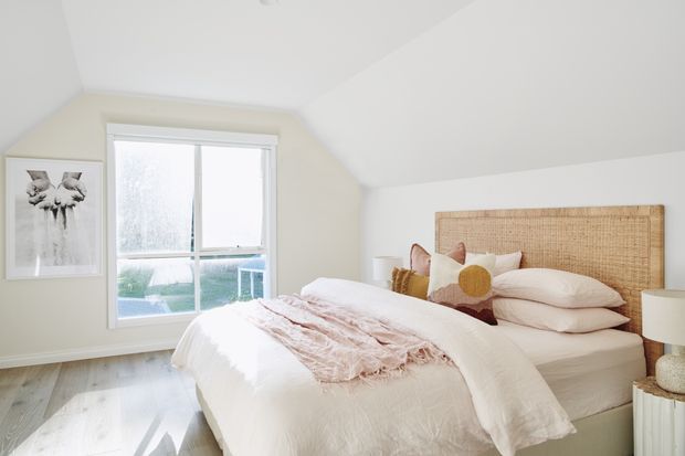 Bright, softly styled bedroom with a sloped ceiling, pale neutral walls, light timber flooring, and a large bed featuring a woven rattan headboard. Layered blush and cream bedding, textured cushions, bedside lamps, and a framed botanical artwork create a warm, relaxed interior.