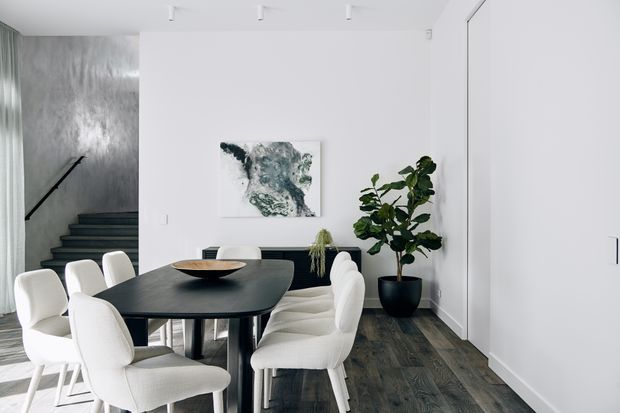 A bright, minimalist dining area with a dark rectangular dining table surrounded by upholstered white chairs. White walls and concealed cabinetry create a clean backdrop, while dark timber flooring, a large indoor plant, abstract artwork, and a stairwell add contrast and visual interest. The space opens toward a stairway at the left.