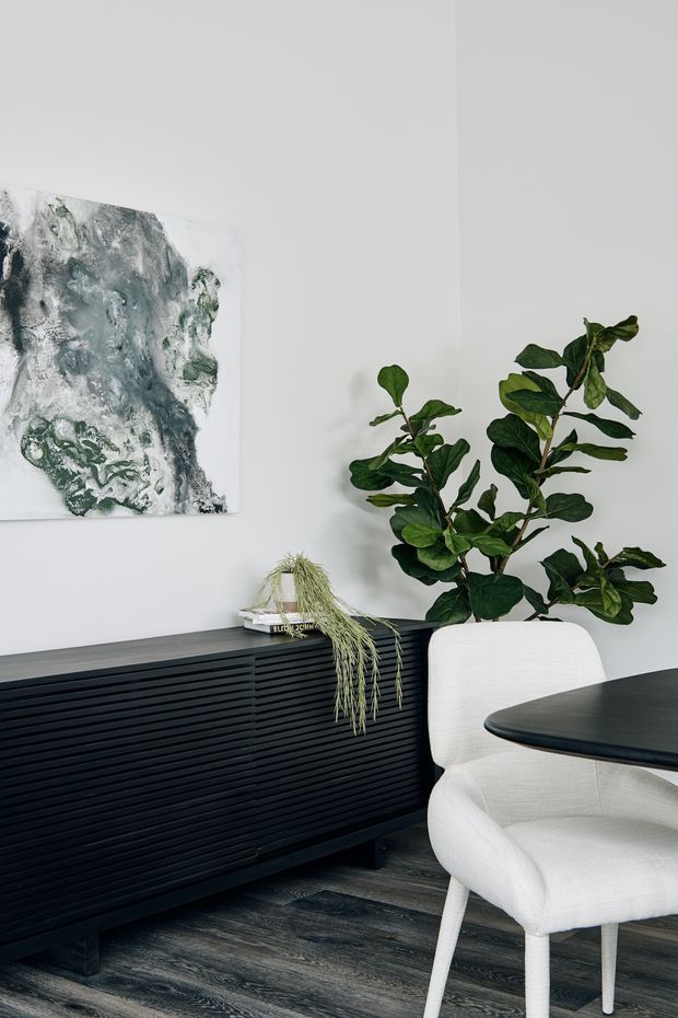 A refined dining area with a white upholstered dining chair and dark rounded tabletop beside a low black fluted sideboard. White walls, dark timber flooring, large leafy indoor plants, and abstract green-grey artwork create a calm, contemporary setting.