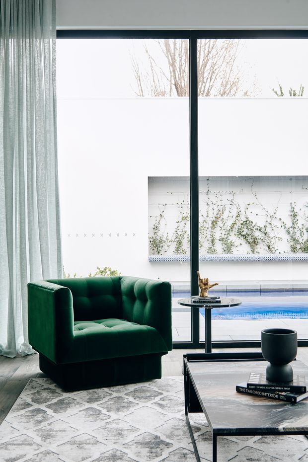 A refined living room with a deep emerald-green tufted armchair beside full-height glazing. Pale sheer curtains frame a black-framed window overlooking a courtyard and swimming pool. A black metal and stone coffee table, sculptural decor, and a patterned grey rug create a sophisticated, high-contrast interior.