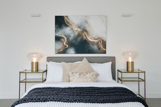 A calm, symmetrical bedroom with a grey upholstered bedhead, white bedding, layered cream and beige pillows, and a dark chunky-knit throw. Matching slim metal-and-glass bedside tables with warm table lamps flank the bed. A blue-grey abstract artwork with metallic accents hangs above, against a light neutral wall.