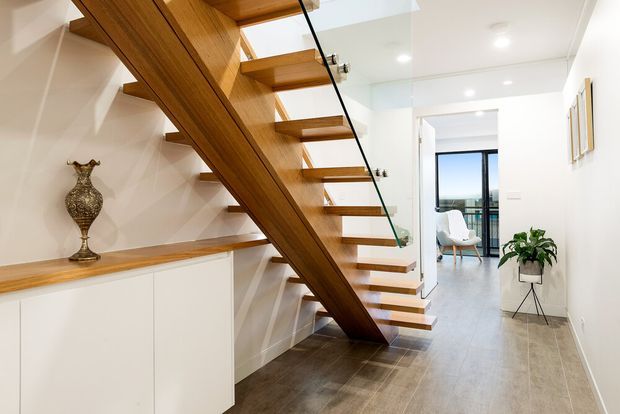 Bright contemporary interior hallway featuring an open-riser floating timber staircase with a substantial diagonal timber stringer and clear glass balustrade. White built-in cabinetry and a timber display ledge run along the left wall, while the tiled floor leads toward a glazed room with a chair and indoor plant in the distance. Recessed ceiling lights and white walls create a clean, airy composition.